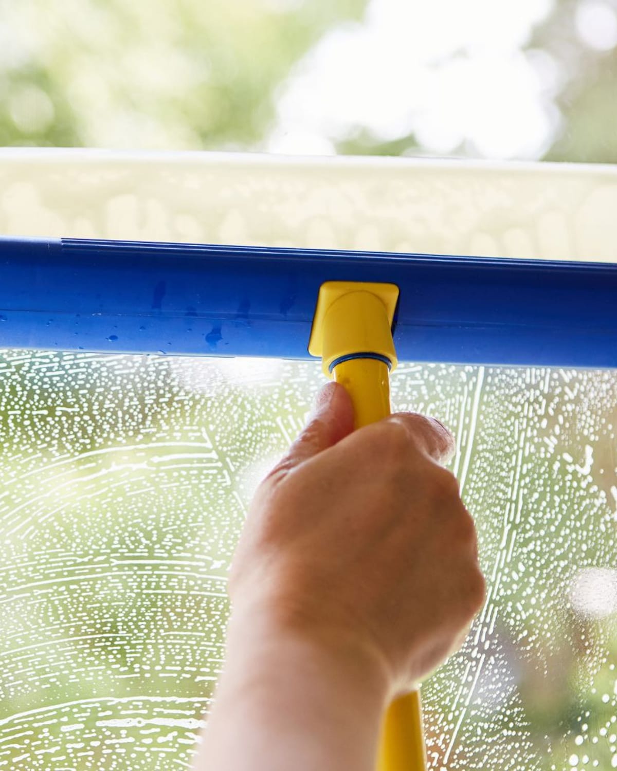 Hand guiding a squeegee across a sudsy windowpane with green trees behind it — everyday work on Centennial, CO glass